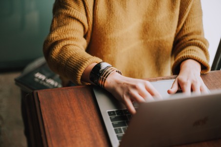 Eine Person, von der nur der Rumpf und die Arme zu sehen sind, sitzt an einem braunen Tisch und arbeitet an einem Laptop. Sie trägt einen beigefarbenen Pullover.