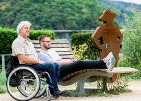 Foto: Zwei Männer sitzen im Freien, einer auf einer Bank, der andere im Rollstuhl