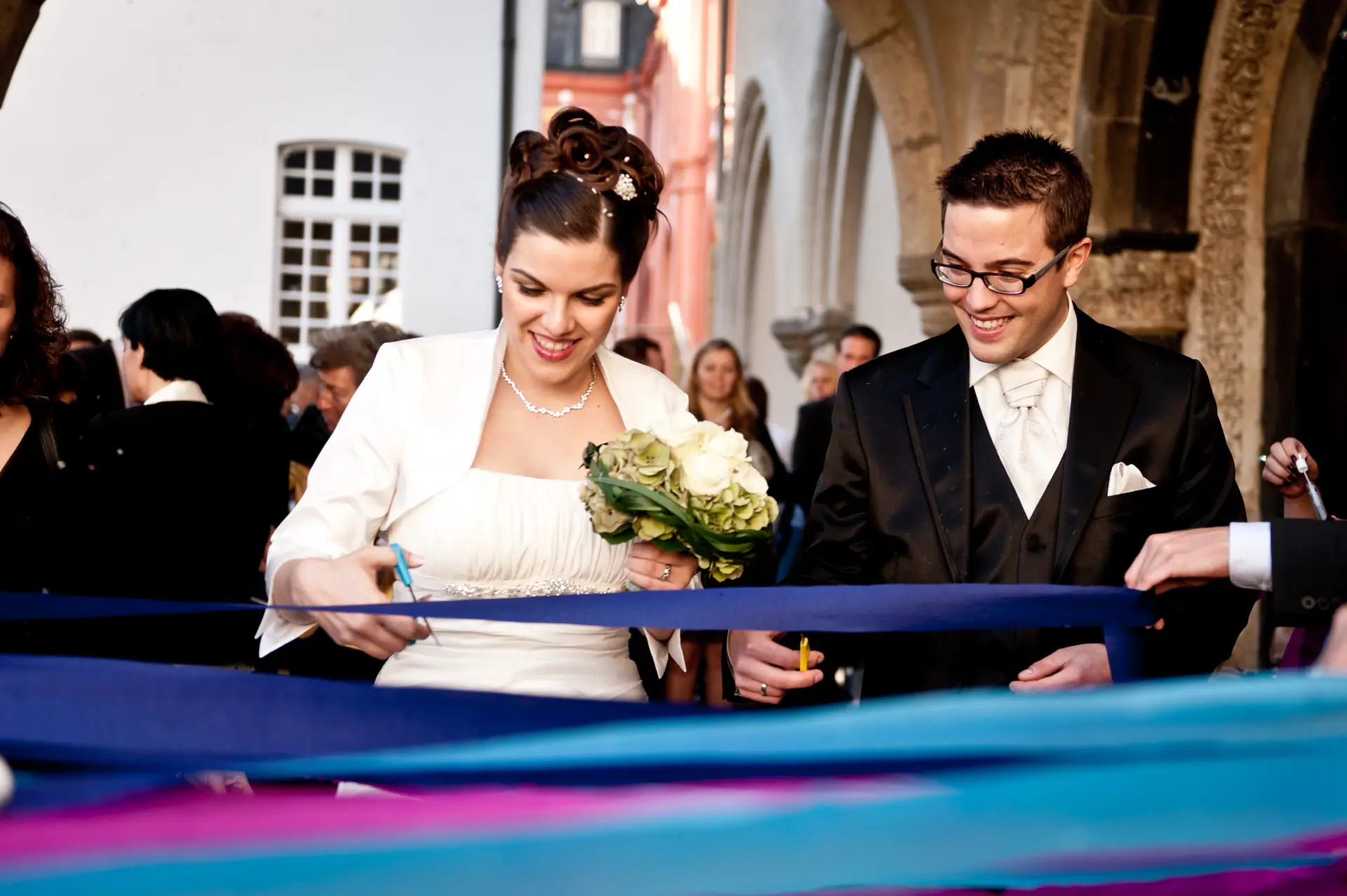 Foto von Hochzeitspaar, das im Marienhof bunte Bänder zerschneidet