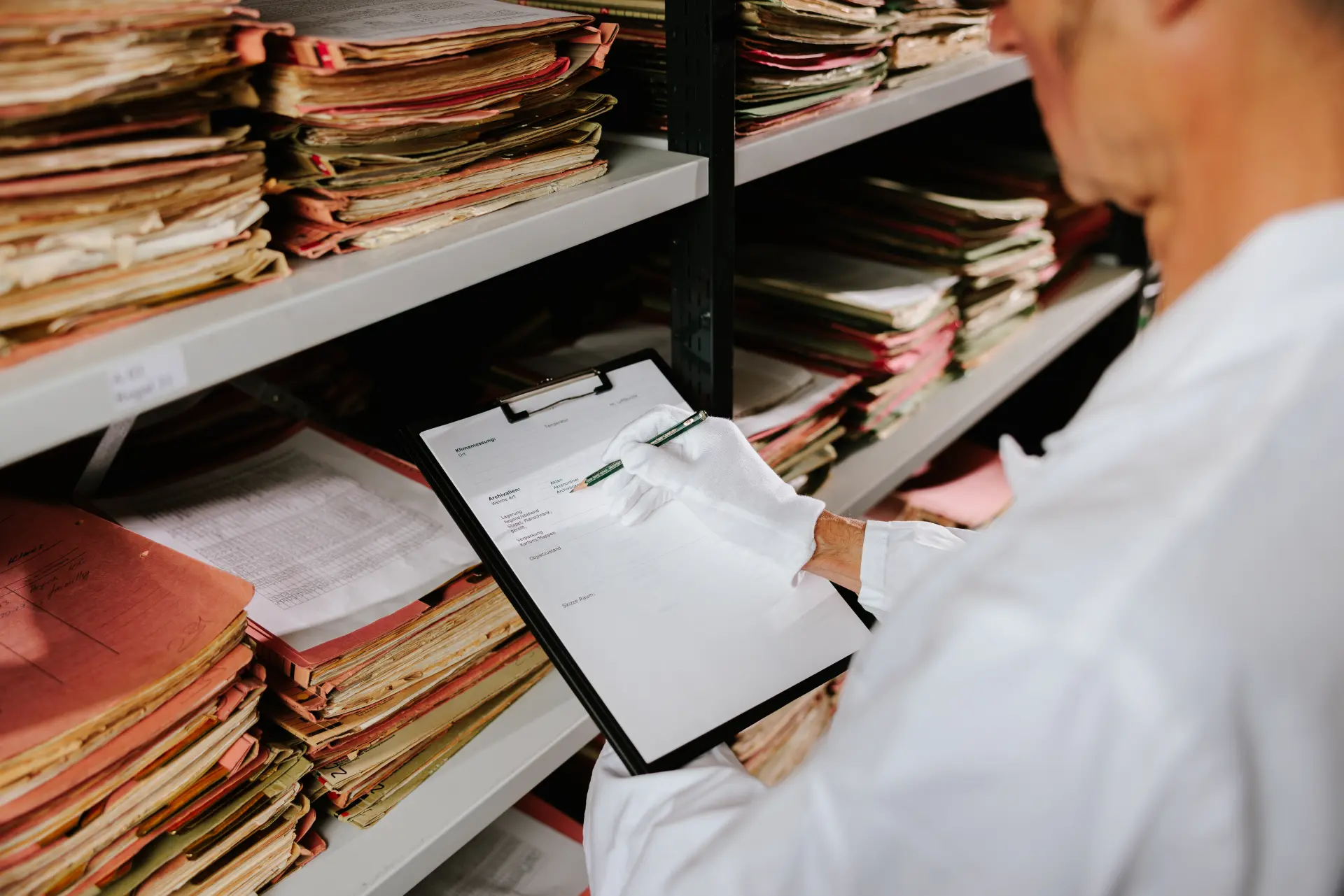 Foto einer Person vor einem Regal mit Aktenmappen, sie hält ein Klemmbrett mit einem Formular in der Hand und füllt dieses aus.