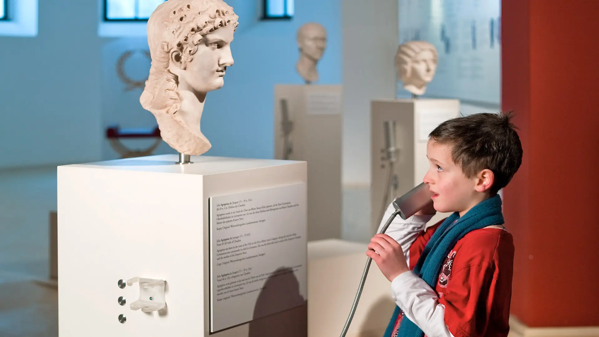 Kind spricht an einer Medienstation scheinbar mit einer Büste der Agrippina