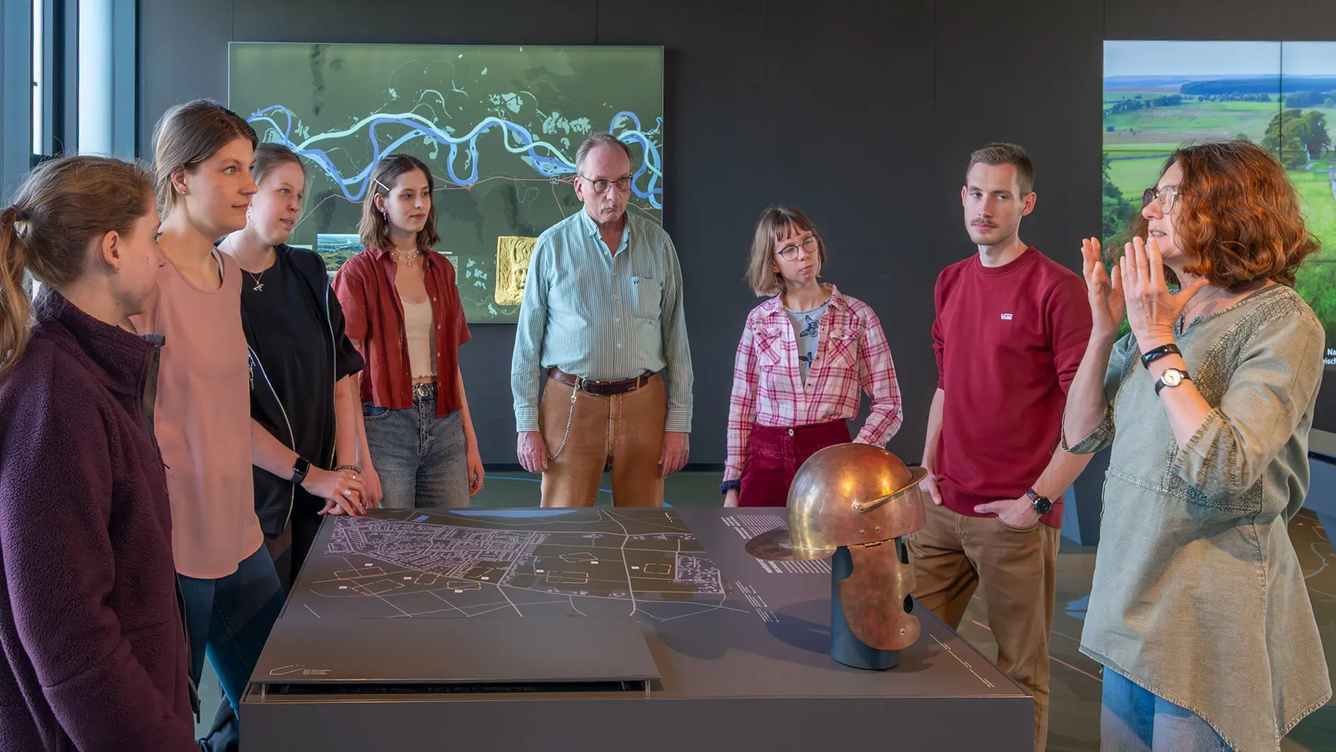 Gruppe von mehreren Erwachsenen um einen Display-Tisch des Limespavillons versammelt, Reiseleiterin in Erklär-Geste