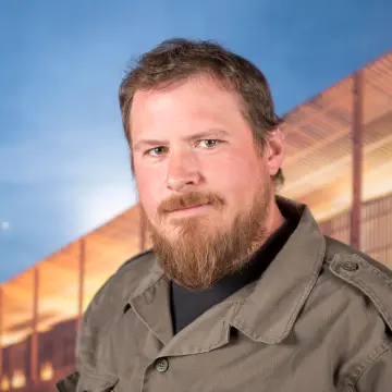 Portraitfoto eines Mannes mit schwarzem Shirt, brauner Jacke und einem langen Bart.