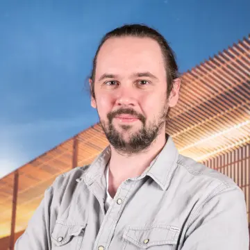 Portraitfoto eines Mannes mit einem Bart, den Haaren zu einem Zopf gebunden und einem grauen Hemd, der lächelnd in die Kamera schaut.