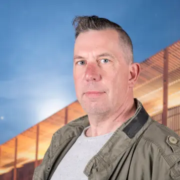 Portraitfoto eines Mannes mit mit kurzen, dunkelgrauen Haaren, beigen Oberteil und grüner Jacke.