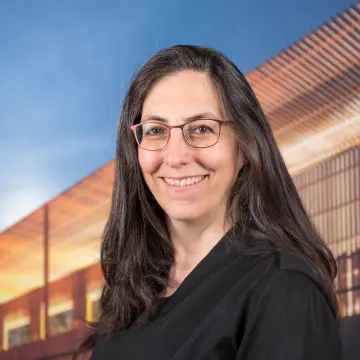 Portraitfoto einer Frau mit schwarzen langen Haaren, einer Brille und einem schwarzen Oberteil, die in die Kamera lächelt.