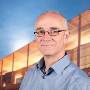 Portraitfoto eines Manne mit Halbglatze, einer Brille und einem blauen Hemd, der lächelnd in die Kamera schaut.