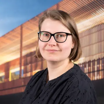 Portraitfoto einer Frau mit kinnlangen, blonden Haaren, einer Brille und einer schwarzen Bluse, die in die Kamera lächelt.