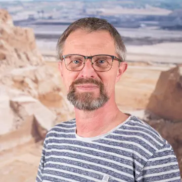 Portraitfoto eines Mannes mit Brille, einem Bart und einem weiß-blau gestreiften Oberteil.