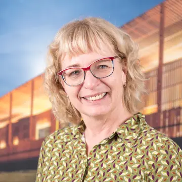 Portraitfoto einer Frau mit blondem, gelocktem Haar, einer Brille und einer gelb-gemusterten Bluse, die in die Kamera lächelt.