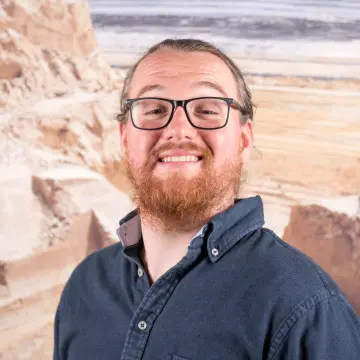Portraitfoto eines Mannes mit einem Bart, den Haaren zu einem Zopf gebunden und einem dunkelblauen Hemd, der lächelnd in die Kamera schaut.