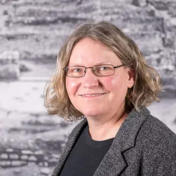Portraitfoto einer Frau mit schulterlangem Haar, einer Brille, einem grauen Sakko und ein schwarzes Shirt, die in die Kamera lächelt.
