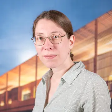 Portraitfoto einer Frau mit Zopf, einer Brille und einer hellen Bluse, die in die Kamera lächelt.