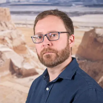 Portraitfoto eines Mannes mit blauem Hemd, einem Bart und einer Brille.