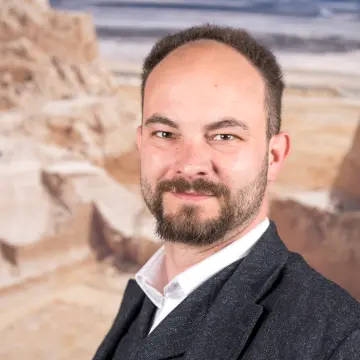 Portraitfoto eines Mannes mit dunklem Haar, einem Bart, einem weißen Hemd und dunkelgrauen Anzug, der lächelnd in die Kamera schaut.