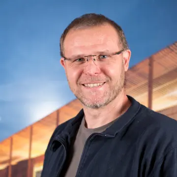 Portraitfoto eines Mannes mit Brille, einem Bart, einem braunem Shirt und einer blauen Jacke, der lächelnd in die Kamera schaut.