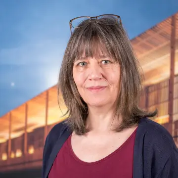 Portraitfoto einer Frau mit schulterlangem Haar und Pony, einem roten Oberteil, einer blauen Jacke und einer Brille auf dem Kopf, lächelt in die Kamera.
