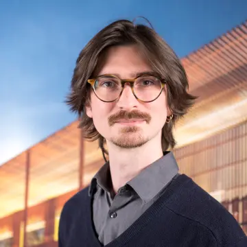 Portraitfoto eines Mannes mit einer Brille, einem Bart, einem grauem Hemd und einem blauen Pullover, lächelt in die Kamera.