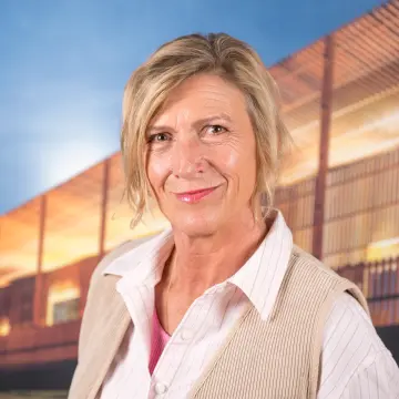 Portraitfoto einer Frau mit blondem Haar, einer hellen Bluse und einer beigen Weste, lächelt in die Kamera.