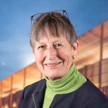 Portraitfoto einer Frau mit kurzem Haar, einer Brille auf dem Kopf, einem grünem Pullover und dunklen Jacke, lächelt in die Kamera. 