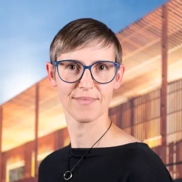 Portraitfoto einer Frau mit einer Brille, kurzem Haar und einem schwarzen Oberteil, die lächelnd in die Kamera schaut.