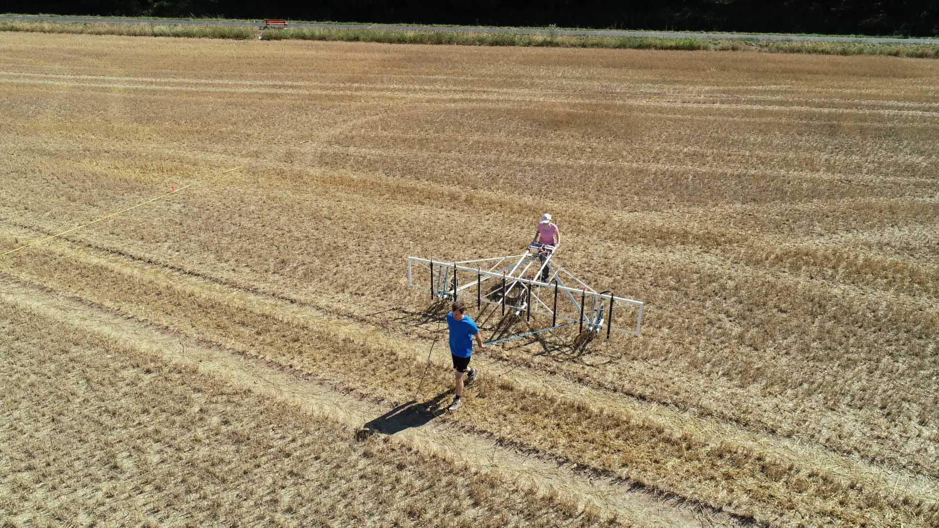 Foto aus der Luft von zwei Personen, die bei sonnigem Wetter eine Messgerät über einen Acker mit gelben Heuresten ziehen