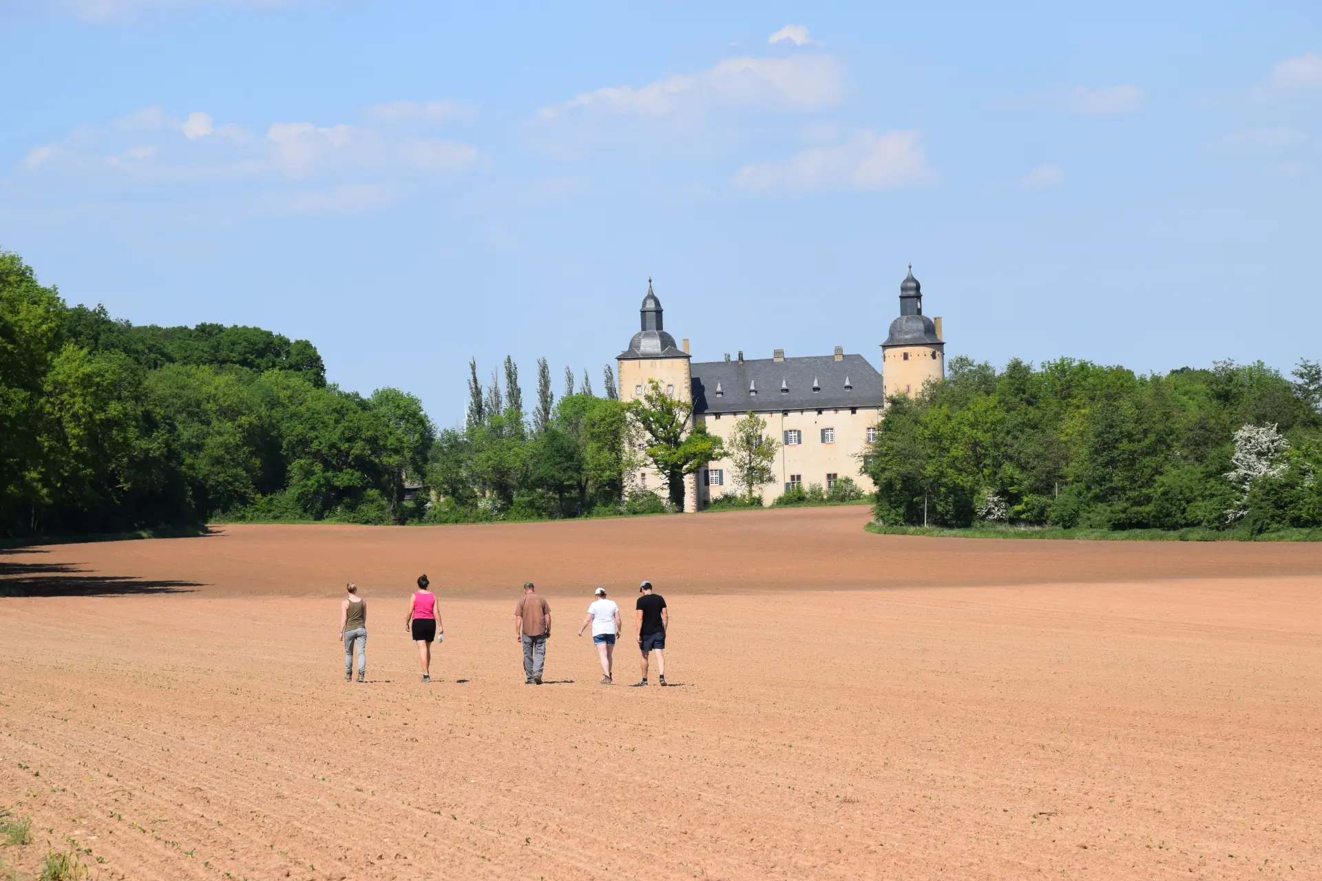 Foto aus der Ferne eines Ackers mit rot-brauner Erde, auf dem fünf Personen mit dem Rücken zur Kamera nebeneinander gehen, im Hintergrund ist eine kleine Burg zu sehen, an beiden Seiten gesäumt von grünen Bäumen.