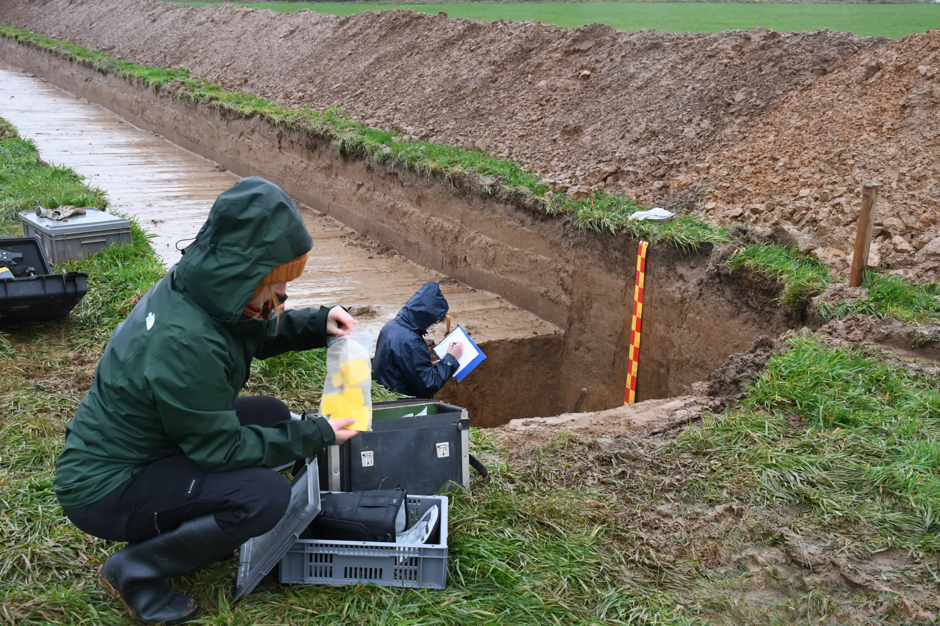 Eine Person in dunkelgrüner Regenjacke sitzt in der Hocke und holt kleine gelbe Schilder aus einer Tüte, im Hintergrund steht eine weitere Person in dunkelblauer Regenjacke in einem quadratischen Loch und notiert etwas auf einem Zeichenbrett.
