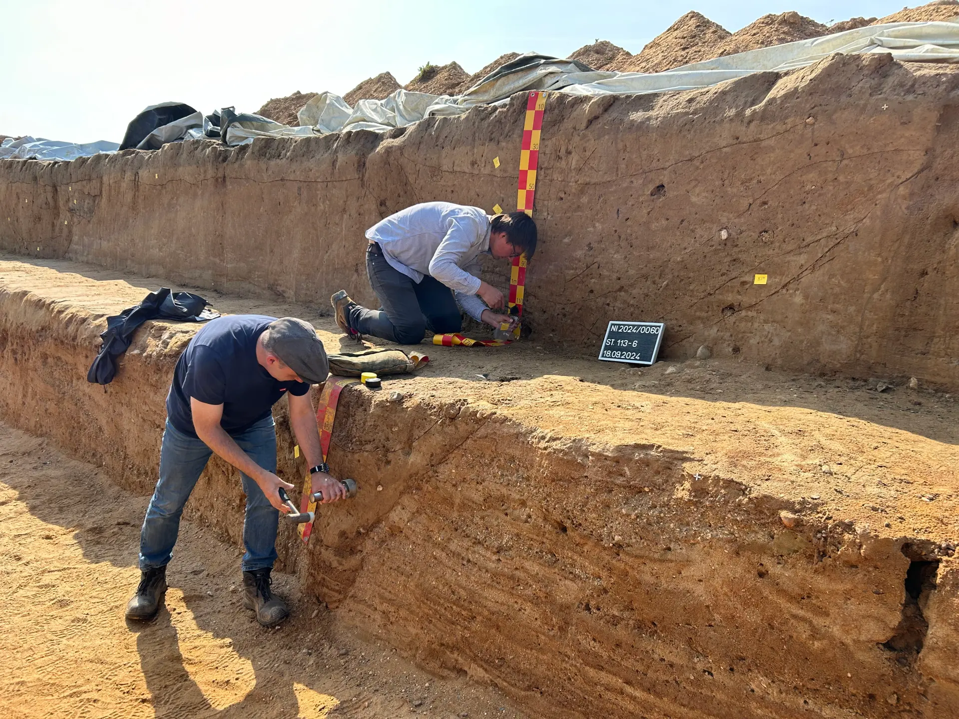 Zwei Personen stehen bei Sonnenschein auf verschiedenen Geländestufen vornübergebeugt in einem Grabungsschnitt mit gelbem Sandboden.