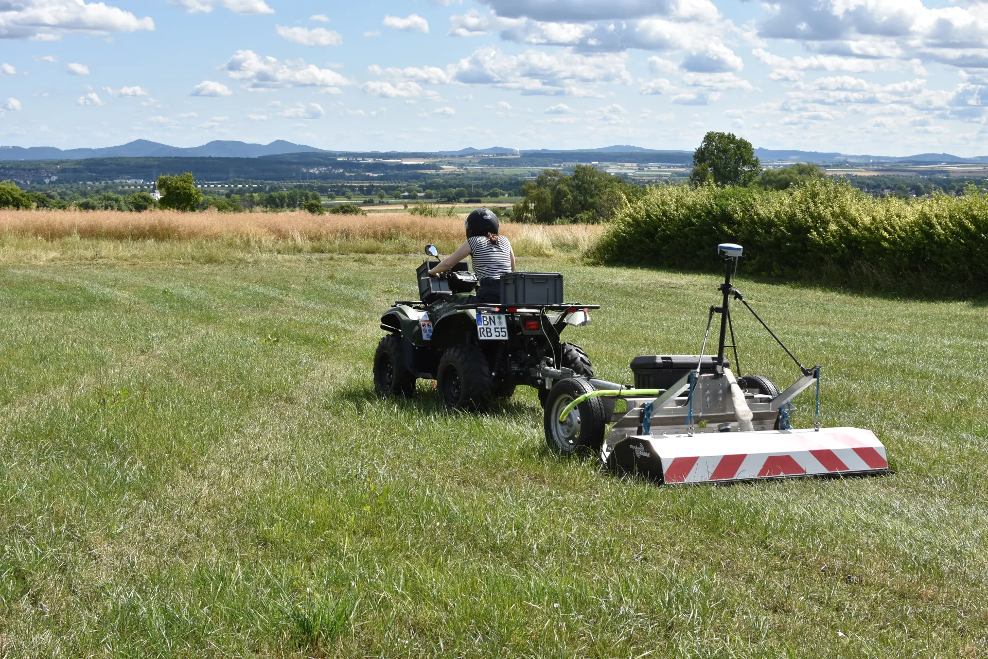 Eine weibliche Person mit einem Motoradhelm auf dem Kopf sitzt auf einem Quad-Bike, das ein Messgerät zieht, und fährt über eine grüne Wiese bei Sonnenschein.