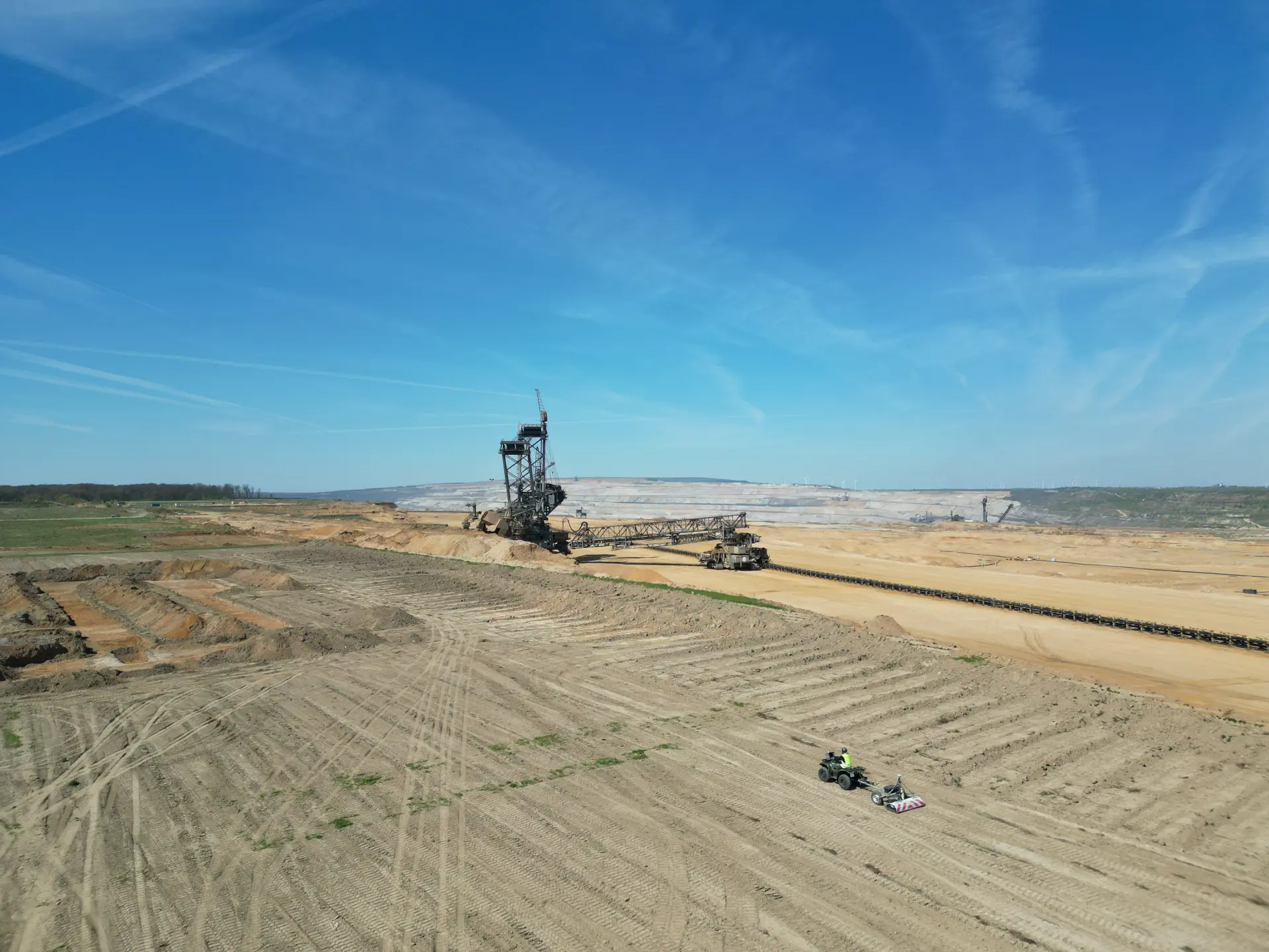 Aufnahme einer gelben, sandigen Fläche aus der Luft bei blauem Himmel mit Blick in den Tagebau, im Hintergrund ein großer Abbaubagger