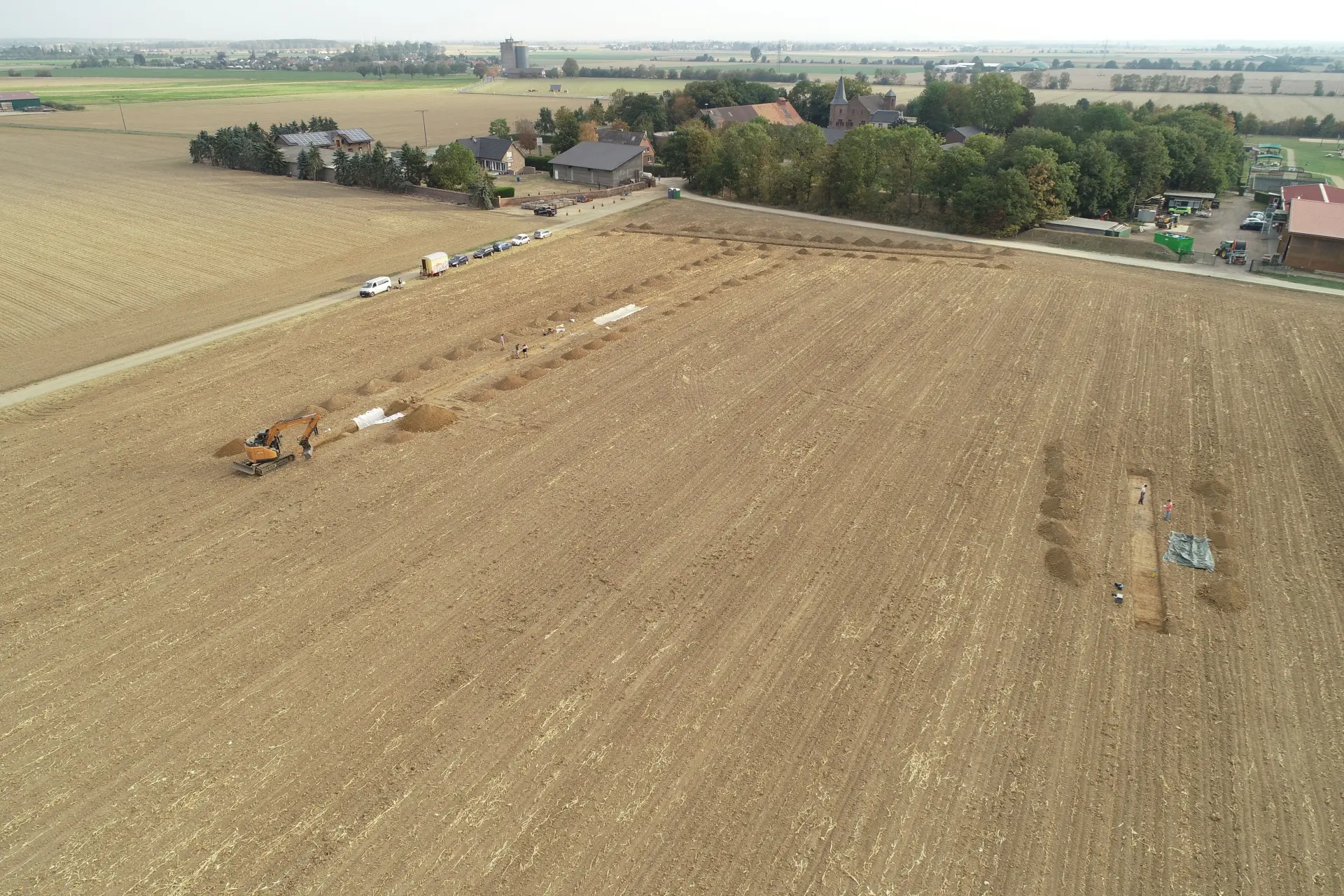 Foto eines hellbraunen Ackers aus der Luft, auf dem mit einem Bagger Grabungsschnitte angelegt werden.