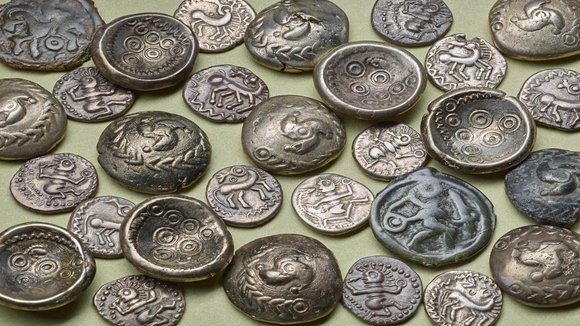 Mehrere antike Silber- und Bronzemünzen liegen verstreut auf hellem Untergrund. Die Münzen zeigen stilisierte Motive wie Pferde, Vögel, menschliche Figuren und kreisförmige Ornamente.
