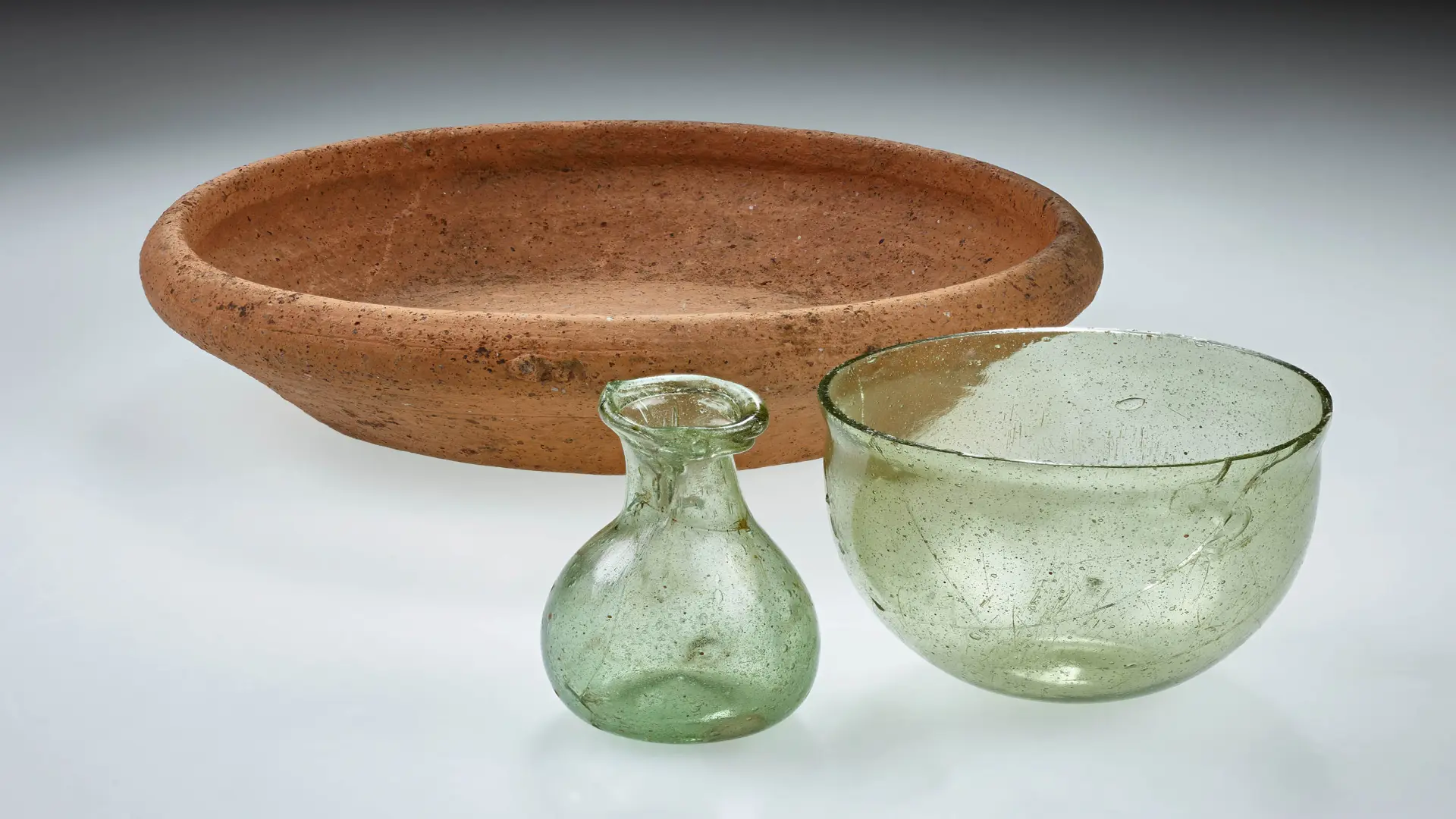 Gefäßbeigaben, darunter ein Glasfläschchen, ein Glasbecher und eine Keramikschüssel aus rotem Ton.