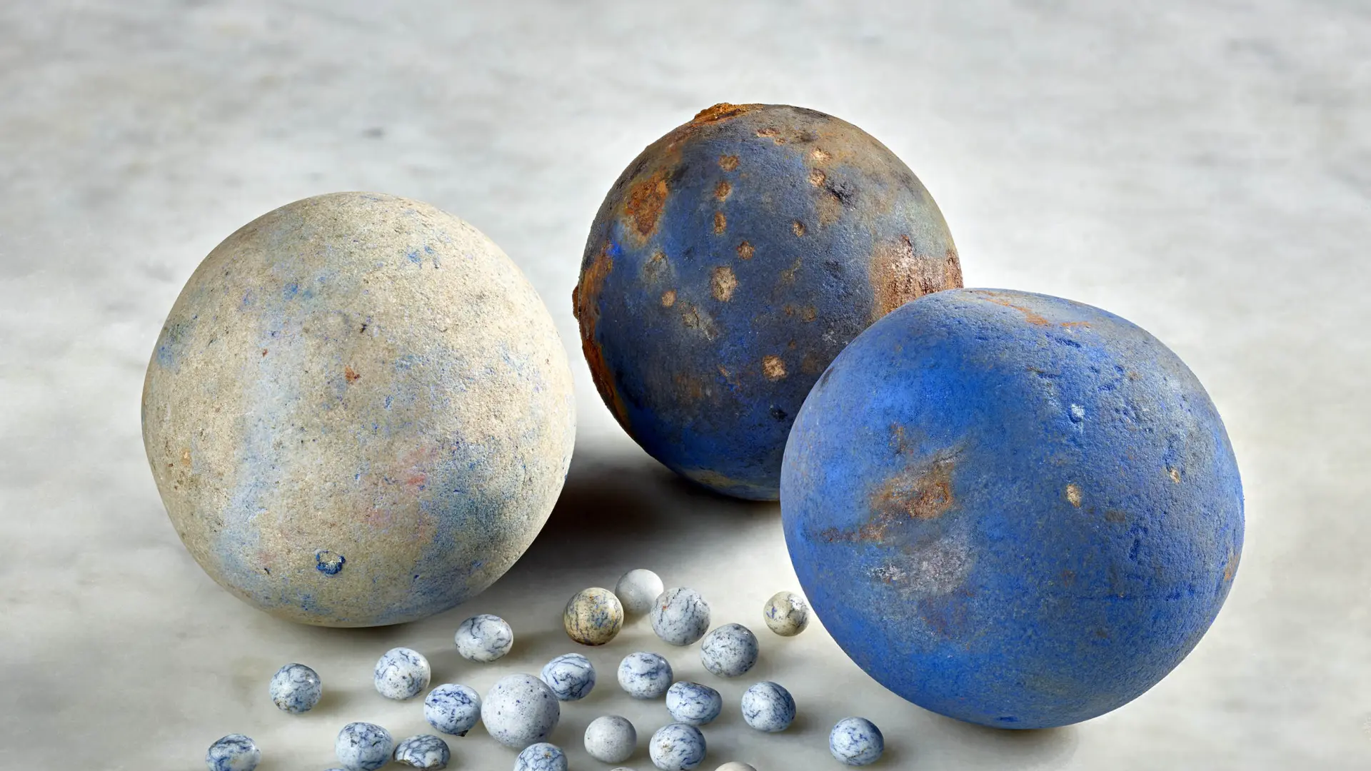 Drei große und zahlreiche kleine Kugeln liegen auf einem hellen Untergrund. Die Oberflächen zeigen unterschiedliche Erhaltungszustände mit blauen Verfärbungen und Gebrauchsspuren.