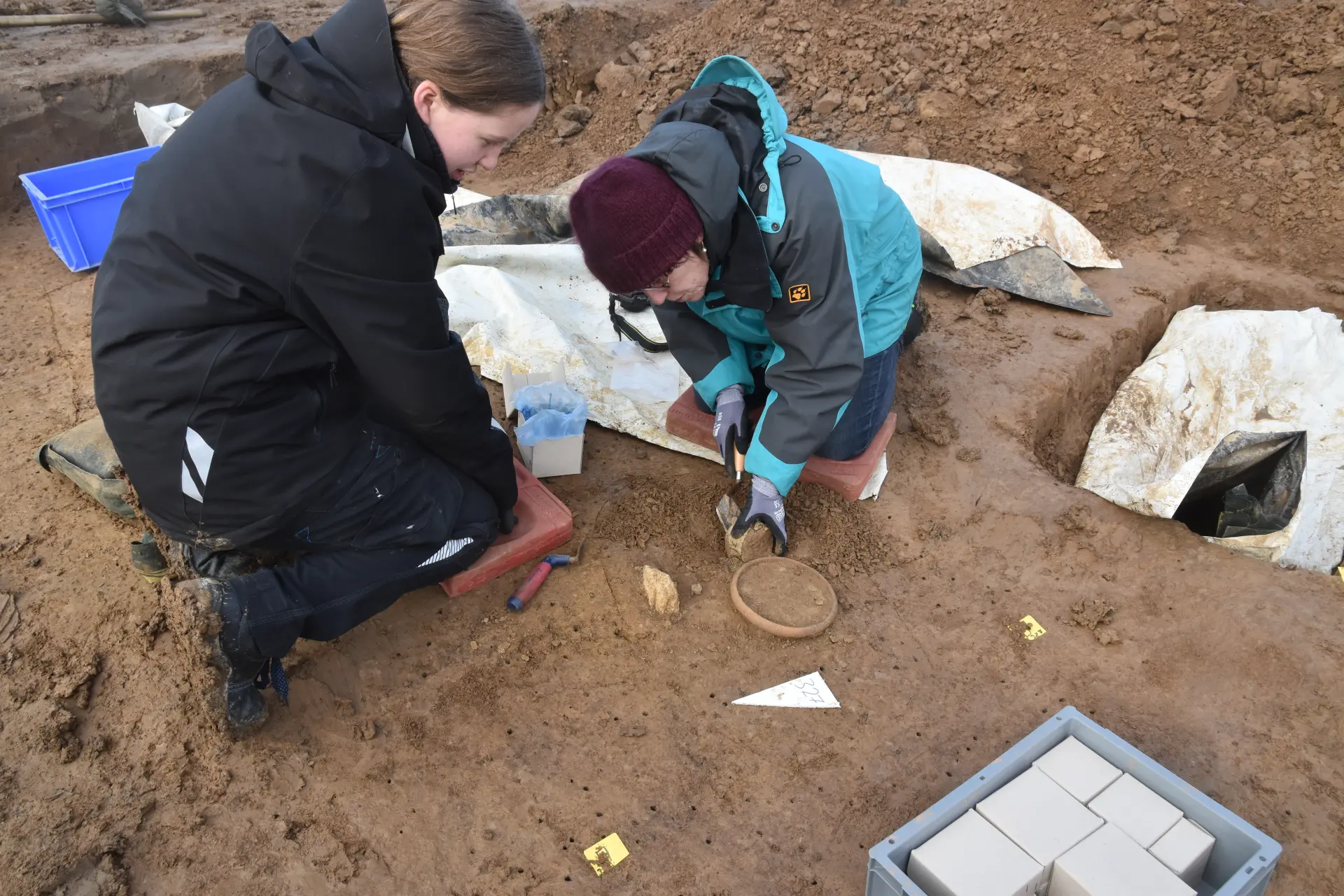 Zwei Archäologinnen arbeiten in einer Ausgrabungsfläche. Mit kleinen Werkzeugen legen sie sorgfältig ein Keramikgefäß im Boden frei.