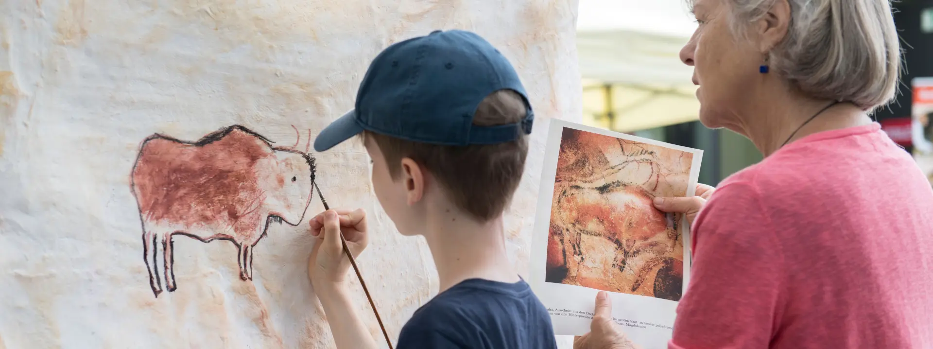 Ein Junge mit Mütze malt einen Bison auf ein an der Wand hängendes Stück Stoff. Eine Person neben ihn hält ein Blatt mit der Zeichnung eines Bisons in der Hand.