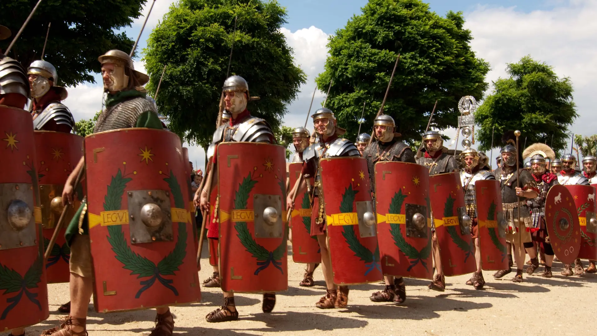 Mehrere männliche Reenactor laufen bei sonnigem Wetter in römischer Rüstung mit roten Schilden in einer Reihe hintereinander