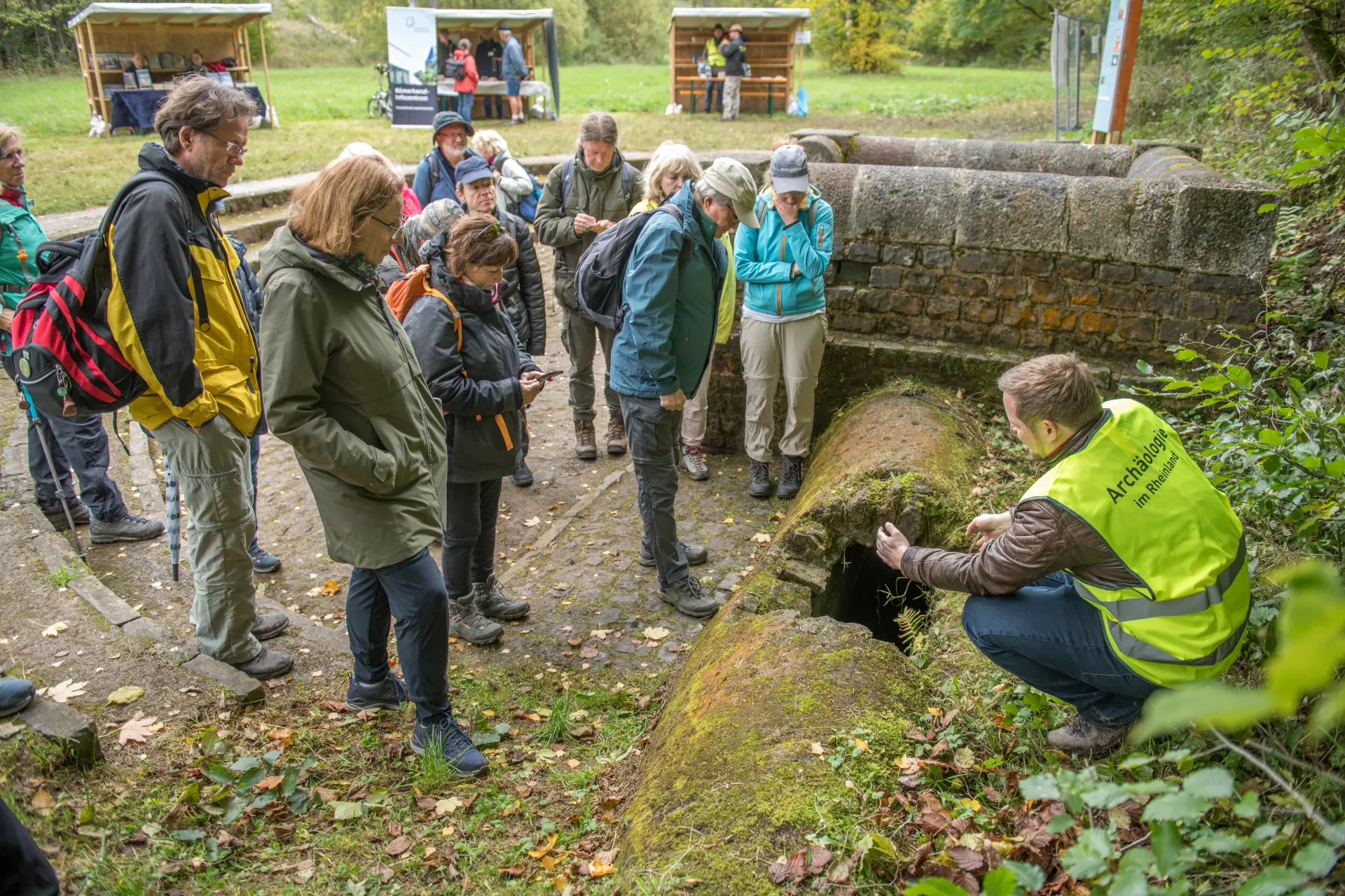 Ein Mann in gelber Warnweste und hockender Position, erklärt einer Personengruppe die römische Wasserleitung. Im Hintergrund sind drei Stände aufgebaut.