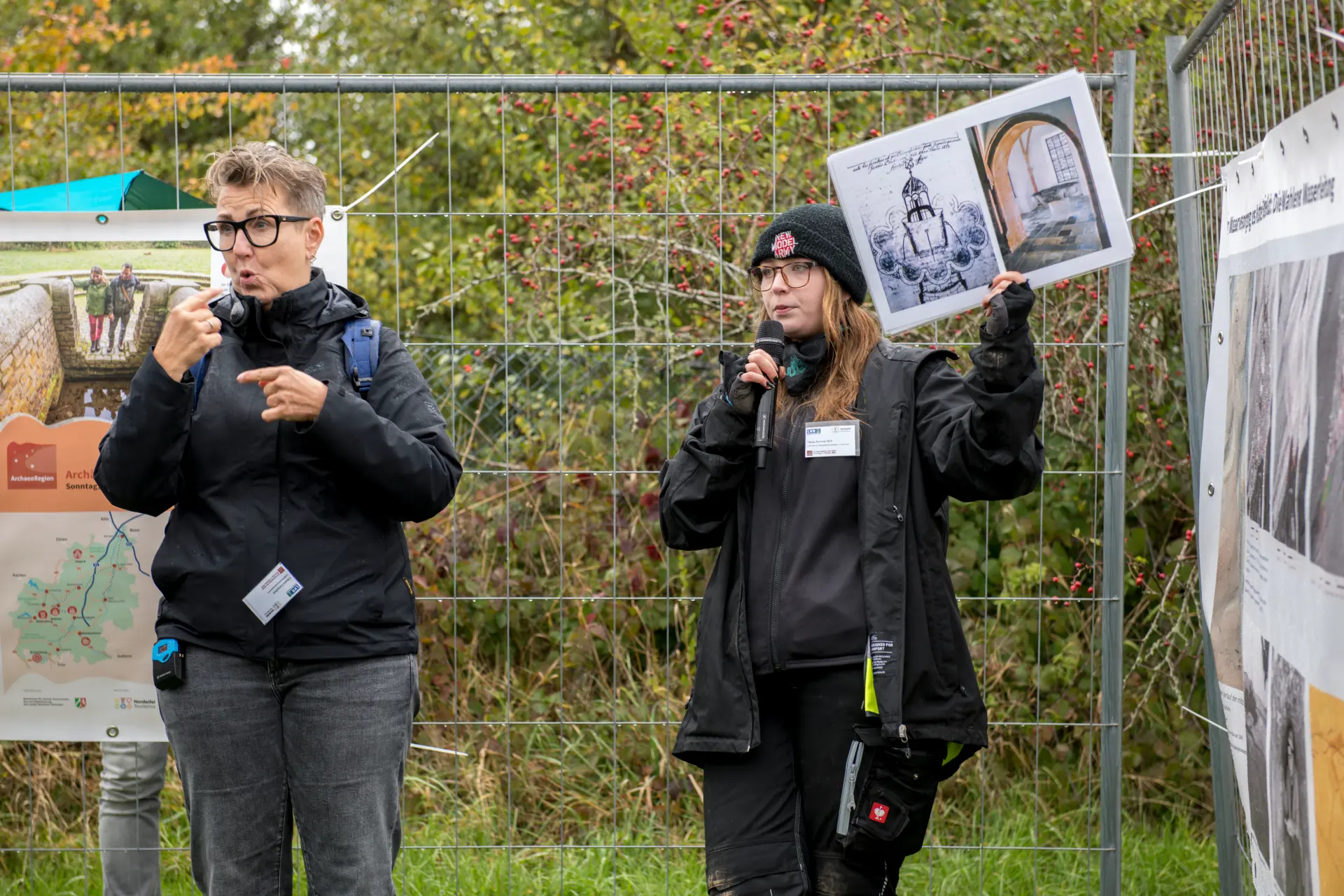 Zwei Frauen stehen vor einem Bauzaun mit einem Plakat. Die linke der beiden übersetzt das gesagte für gehörlose Personen. Die rechte Frau hält ein Informationszettel mit Bildern in die Luft und spricht in ein Mikrofon.