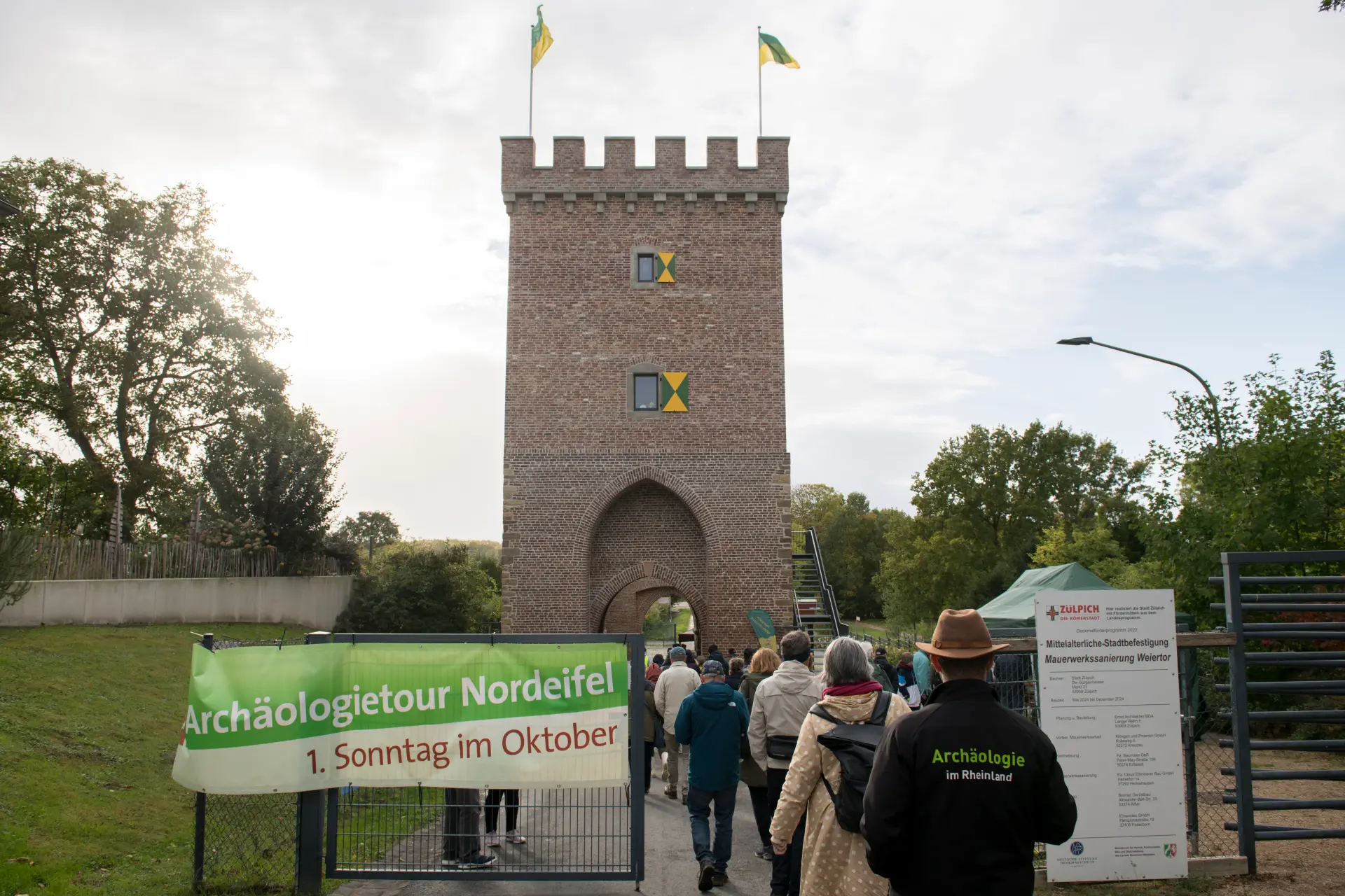 Eine Gruppe von Personen läuft auf einen Turm zu. An einem Zaun hängt ein Plakat von der Archäologietour Nordeifel.