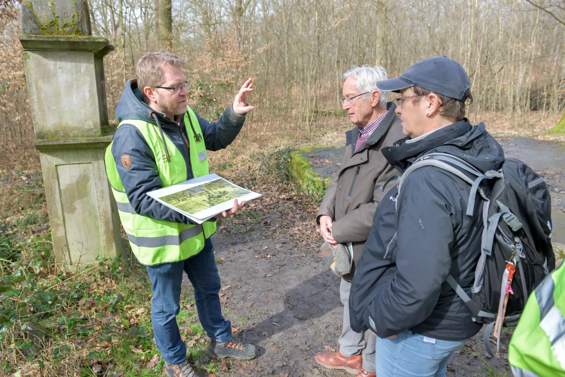 Ein Mann mit gelber Warnweste hält ein Blatt in der Hand und gestikuliert mit seiner anderen Hand. Er erklärt den zwei Personen, die vor ihm stehen etwas.