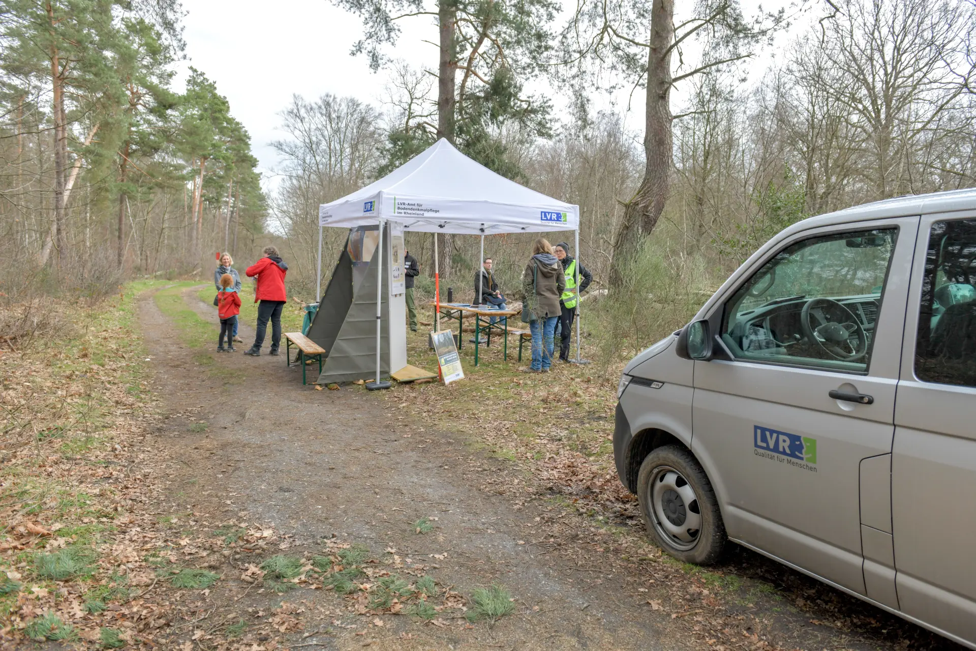 Rechts im Bild steht ein Auto. Dahinter ist ein weißer Pavillon aufgebaut, der als Informationsstand dient. Links ist ein Wald.