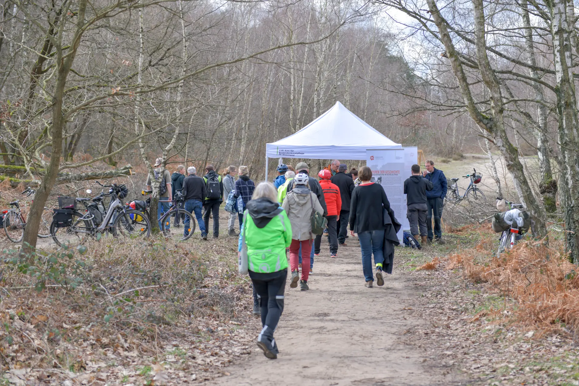 Im Wald steht ein weißer Pavillon und darum stehen Personen und Fahrräder. Einige Personen laufen auf einem Weg darauf zu.