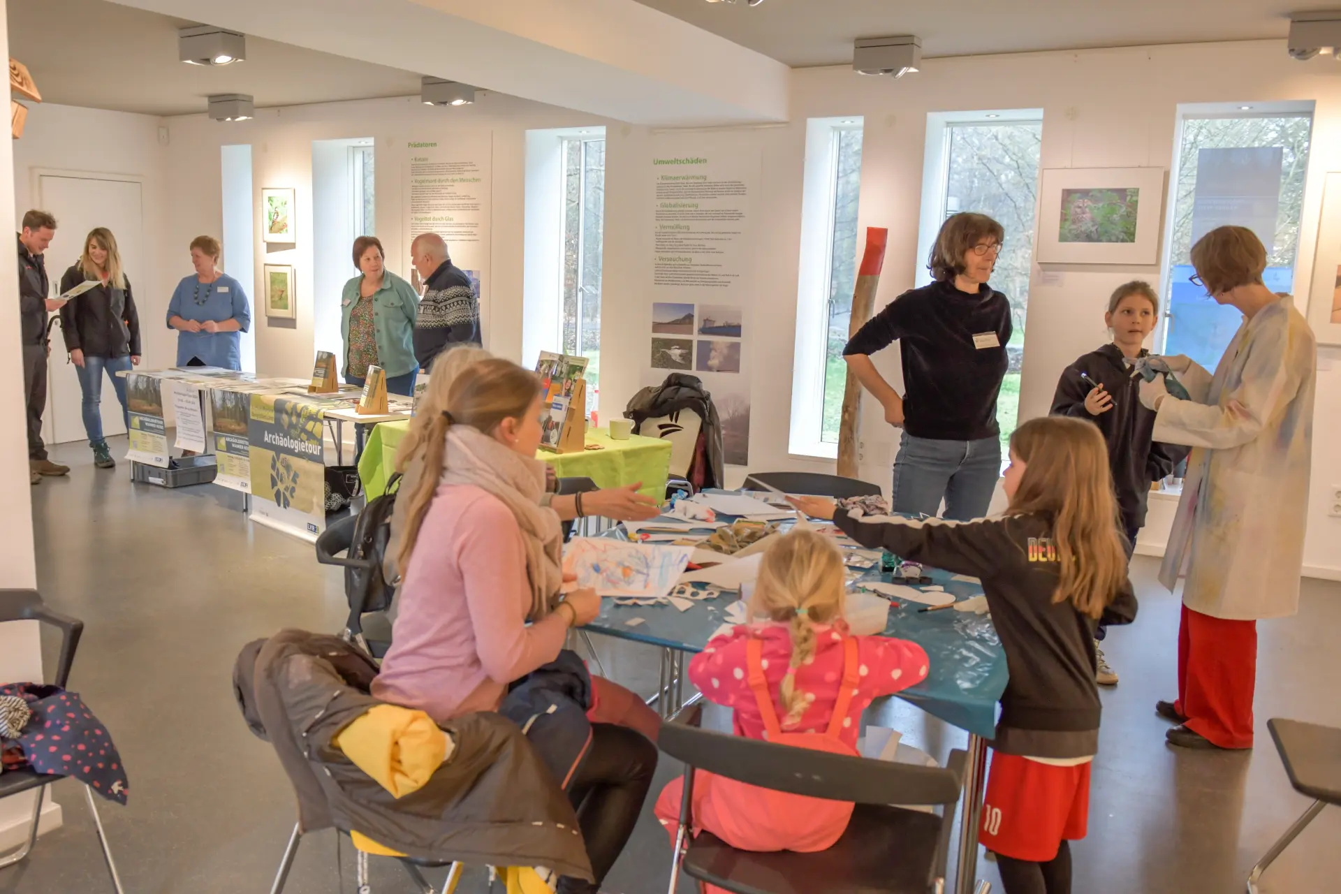 Rechts im Vordergrund steht ein Tisch mit Stühlen an dem Kinder sitzen und basteln. Im Hintergrund ist ein Infostand aufgebaut um den fünf Personen stehen.