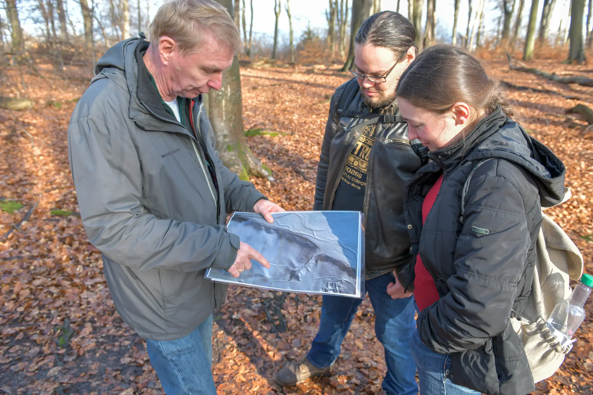 Drei Personen stehen im Wald. Ein Mann zeigt mit seinem Finger auf ein Blatt und erklärt den anderen zwei Personen etwas.