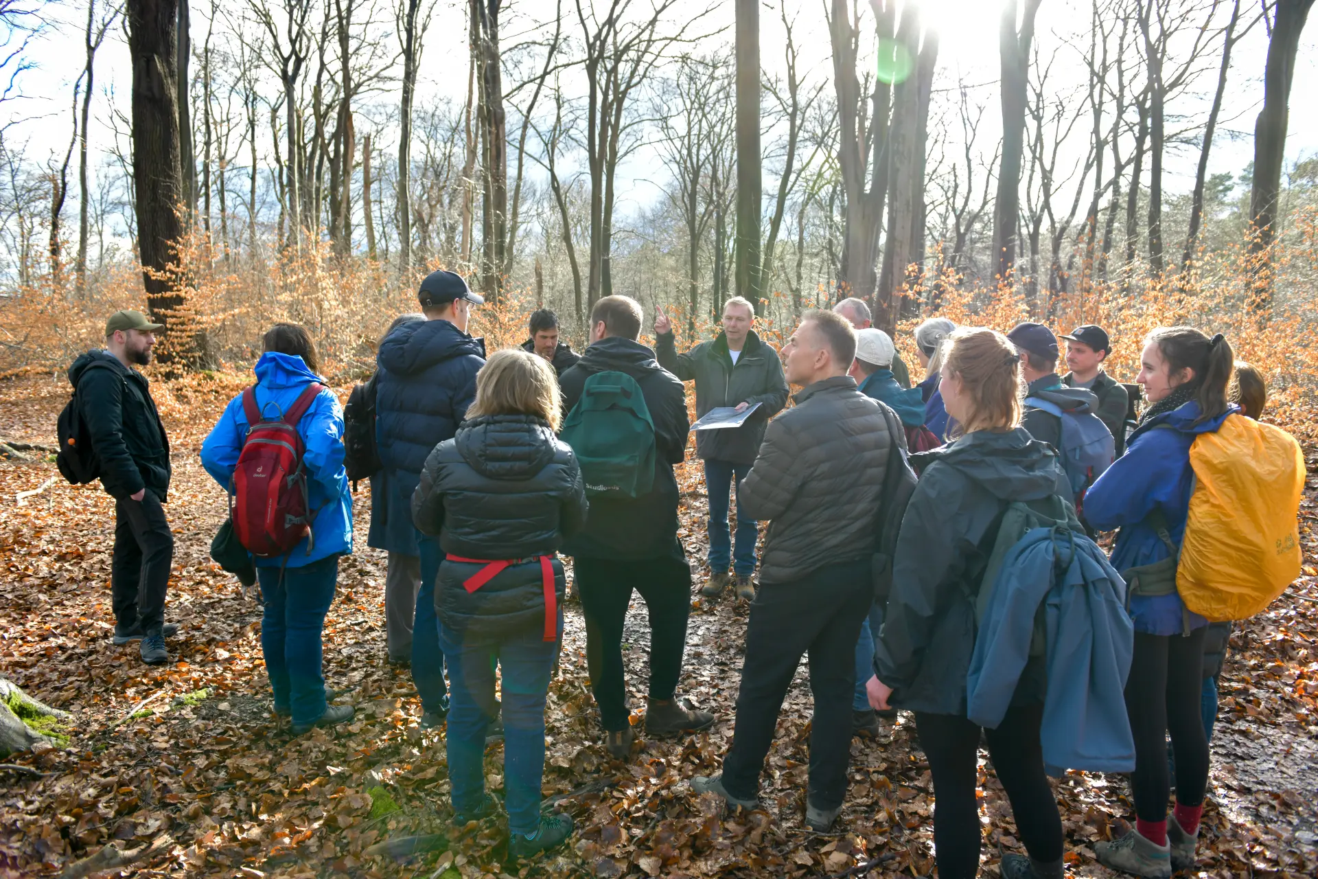Eine Gruppe von Personen steht im Wald. Vor der Gruppe steht ein Mann mit einem Papier in der Hand. Mit der anderen Hand zeigt er auf etwas rechts von ihm.