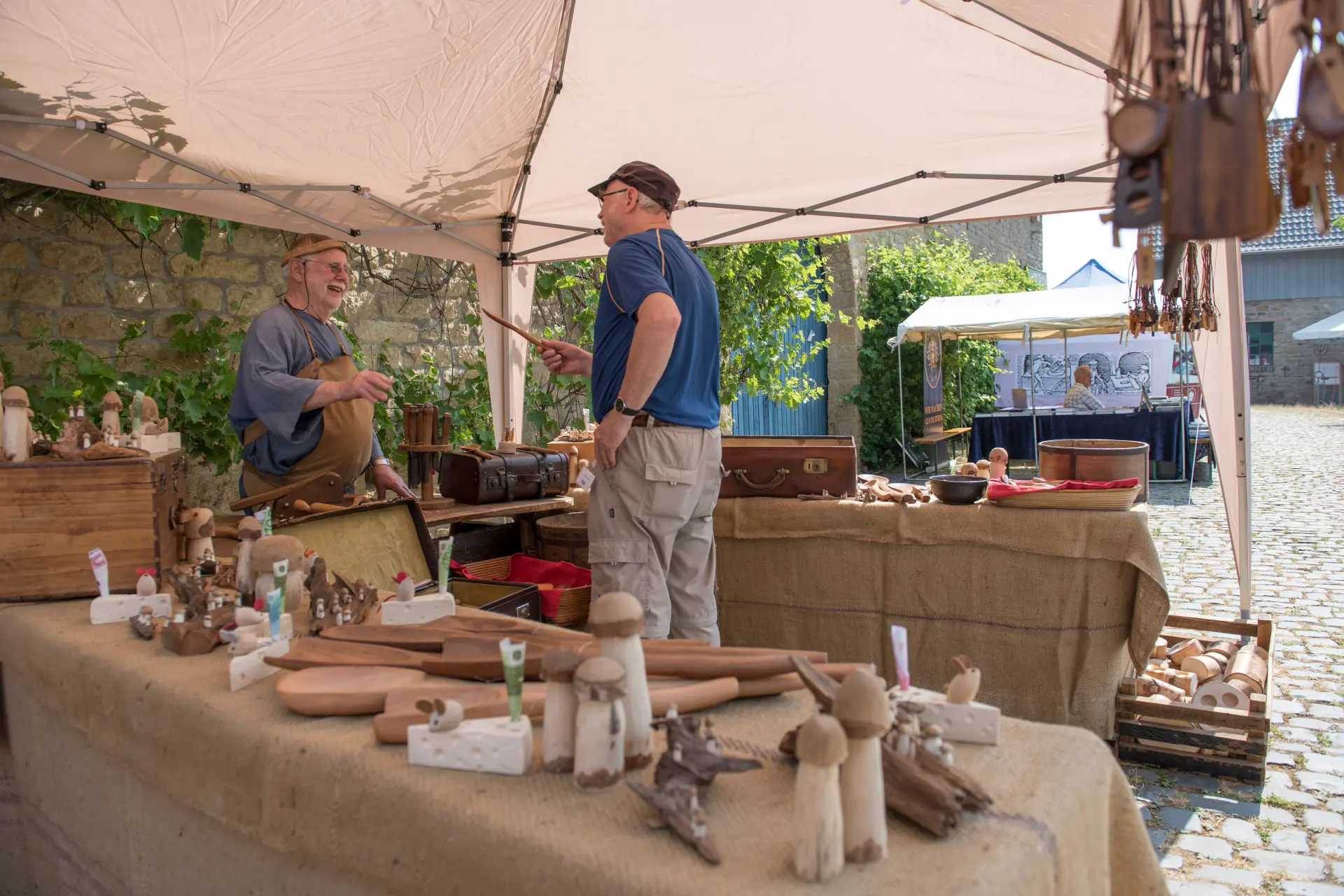 Zwei Männer unter einem Pavillon unterhalten sich. Es ist ein Stand aufgebaut und einer der Männer verkauft dort Holzwaren.