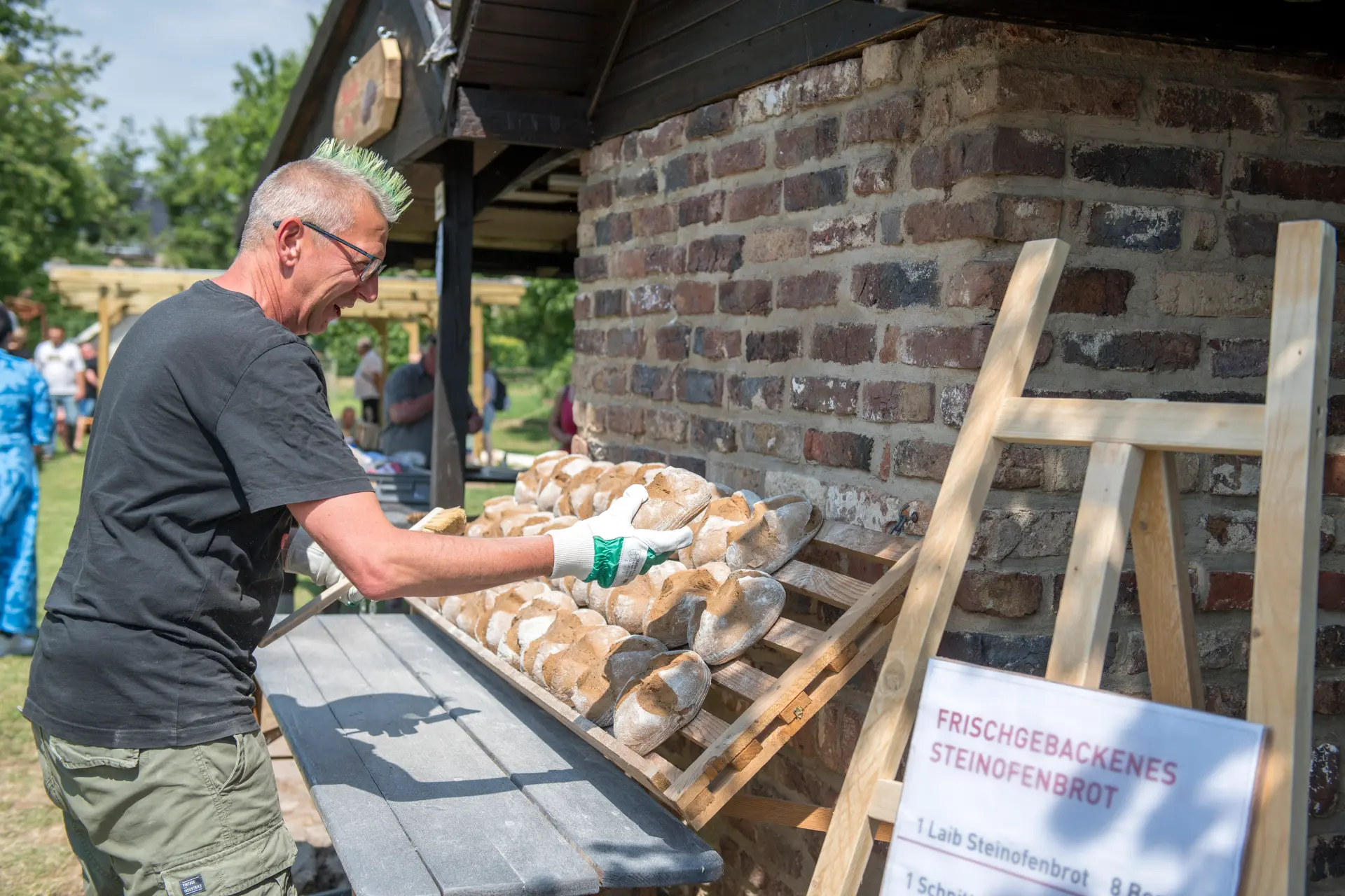 Vor einem Backsteinhaus legt ein Mann Brote auf eine Auslege. Im Vordergrund ist ein Zettel mit der Aufschrift: Frischgebackenes Steinofenbrot.