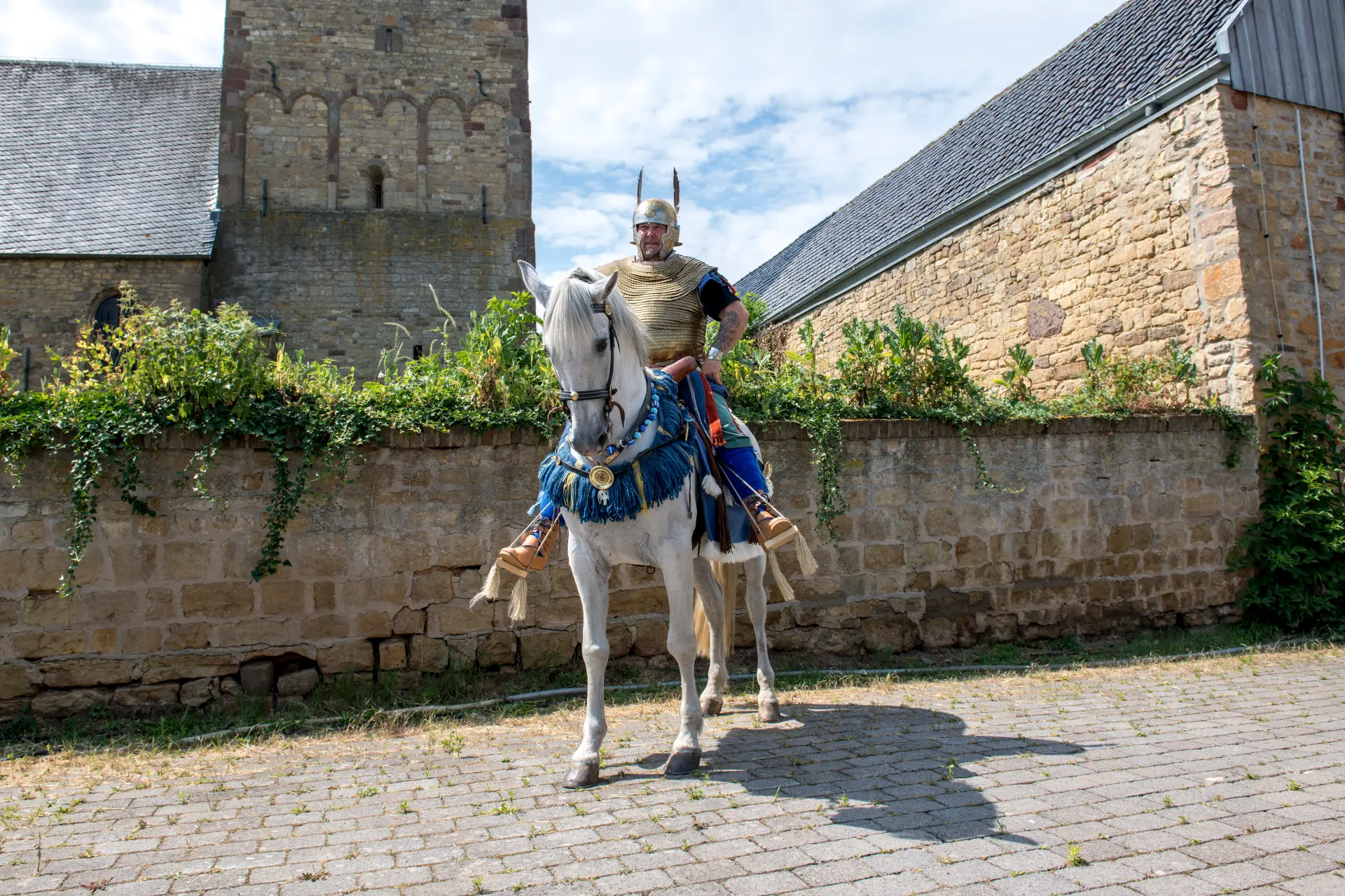 Ein Mann in einer römischen Uniform sitz auf einem weißen Pferd. Er steht vor einer Mauer, dahinter liegen Gebäude und eine Kirche.